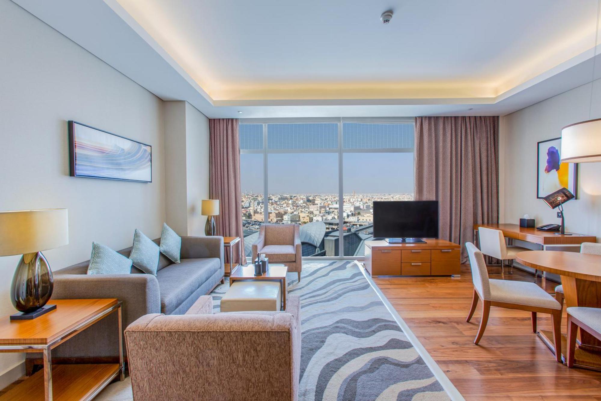 Hilton Riyadh Hotel & Residences Kültér fotó The photo shows a modern hotel room or suite. The space features a stylish living area with a sofa and armchairs arranged around a coffee table. There are lamps providing ambient lighting, and a large window offers a view of the cityscape outside. A 
