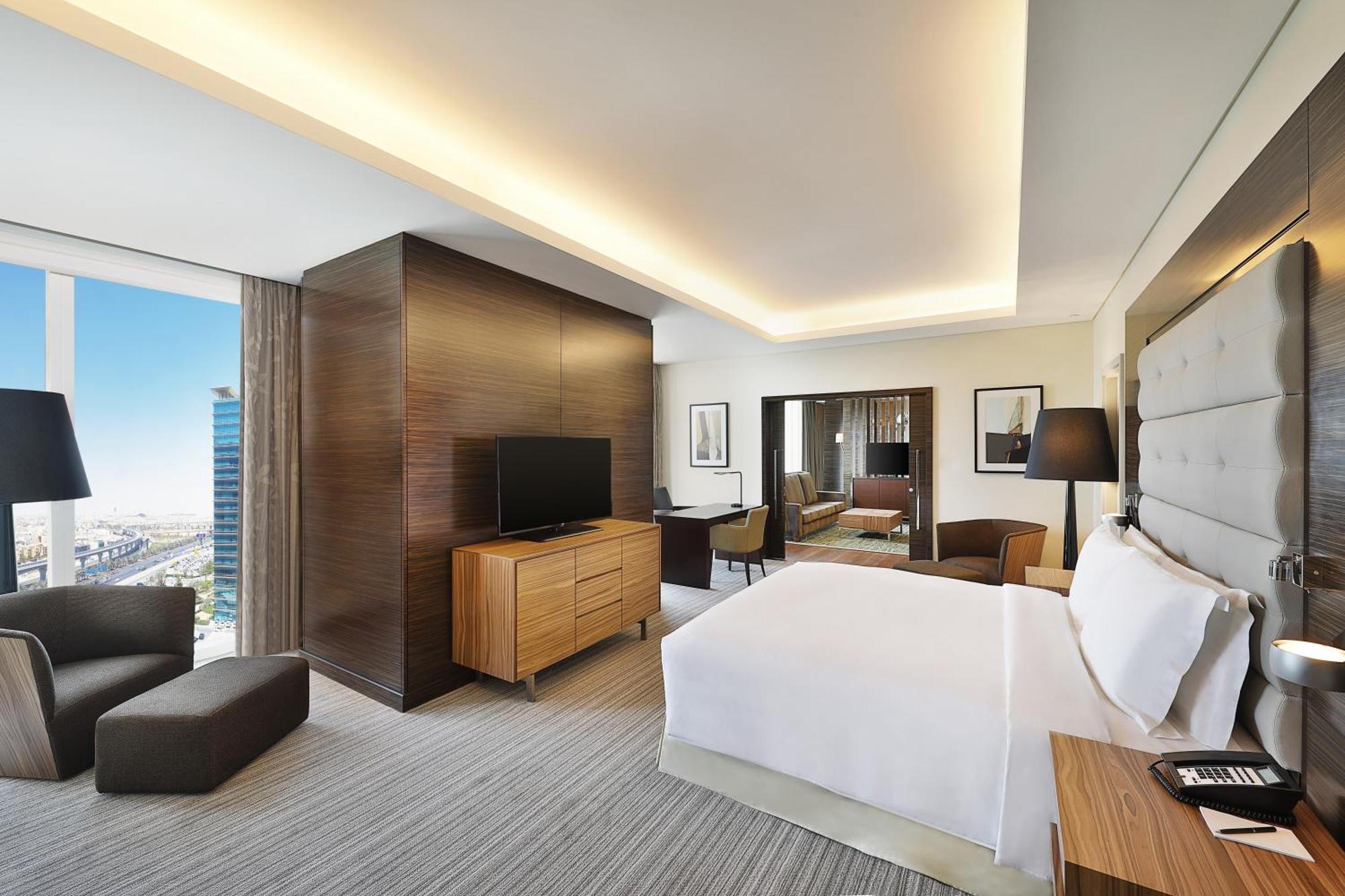 Hilton Riyadh Hotel & Residences Kültér fotó The photo shows a modern hotel room. It features a neatly made bed with white linens, a wooden nightstand beside it, and a cozy armchair. The room has a large window that provides a view of the outside, likely showcasing a cityscape. A flat-screen TV