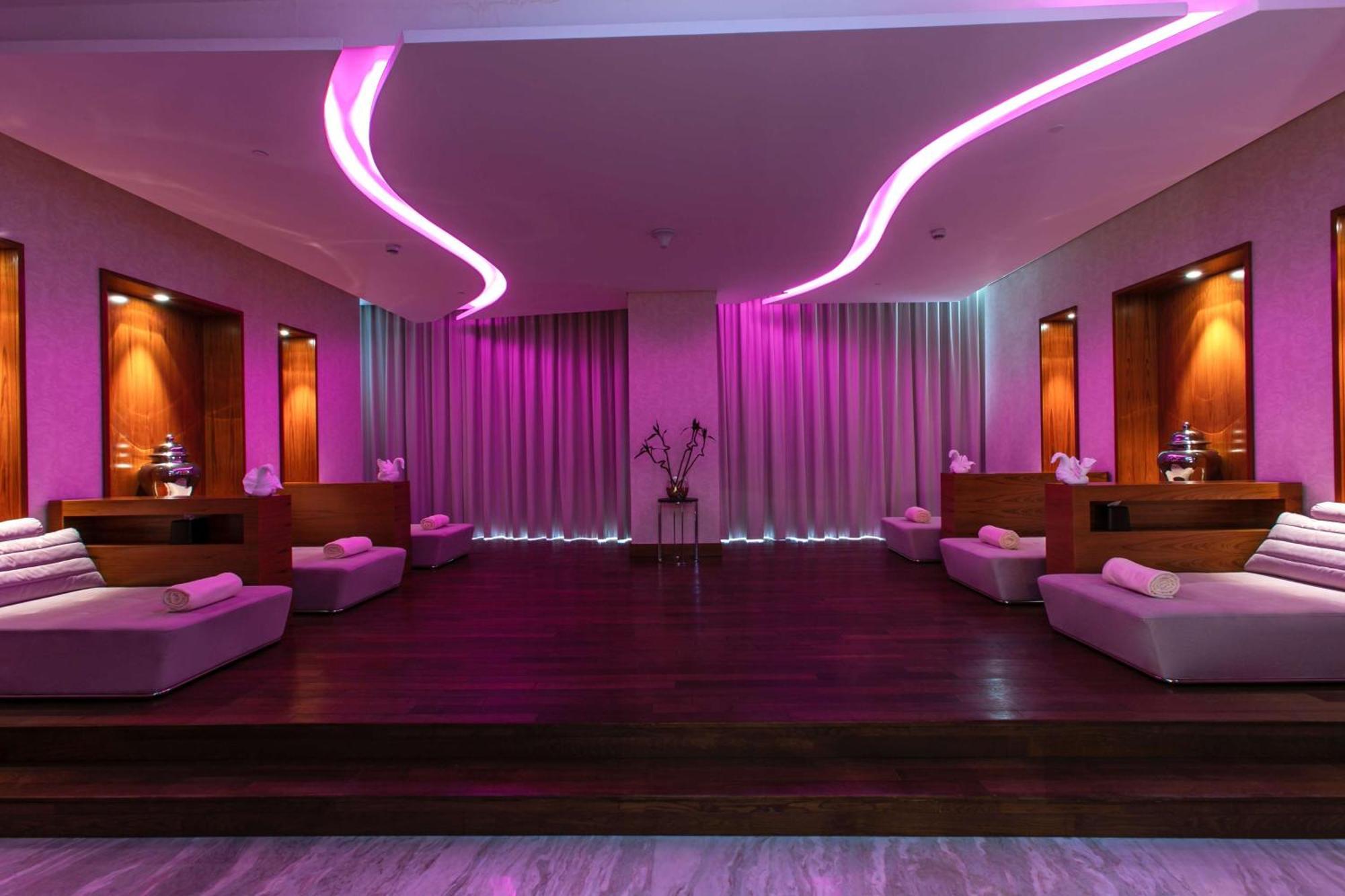 Hilton Riyadh Hotel & Residences Kültér fotó The photo depicts a relaxing interior space, likely a spa or wellness area. It features a wooden floor with a raised platform accented by soft, low couches on either side. The walls are adorned with modern lighting, which emits a calming pink hue. Th