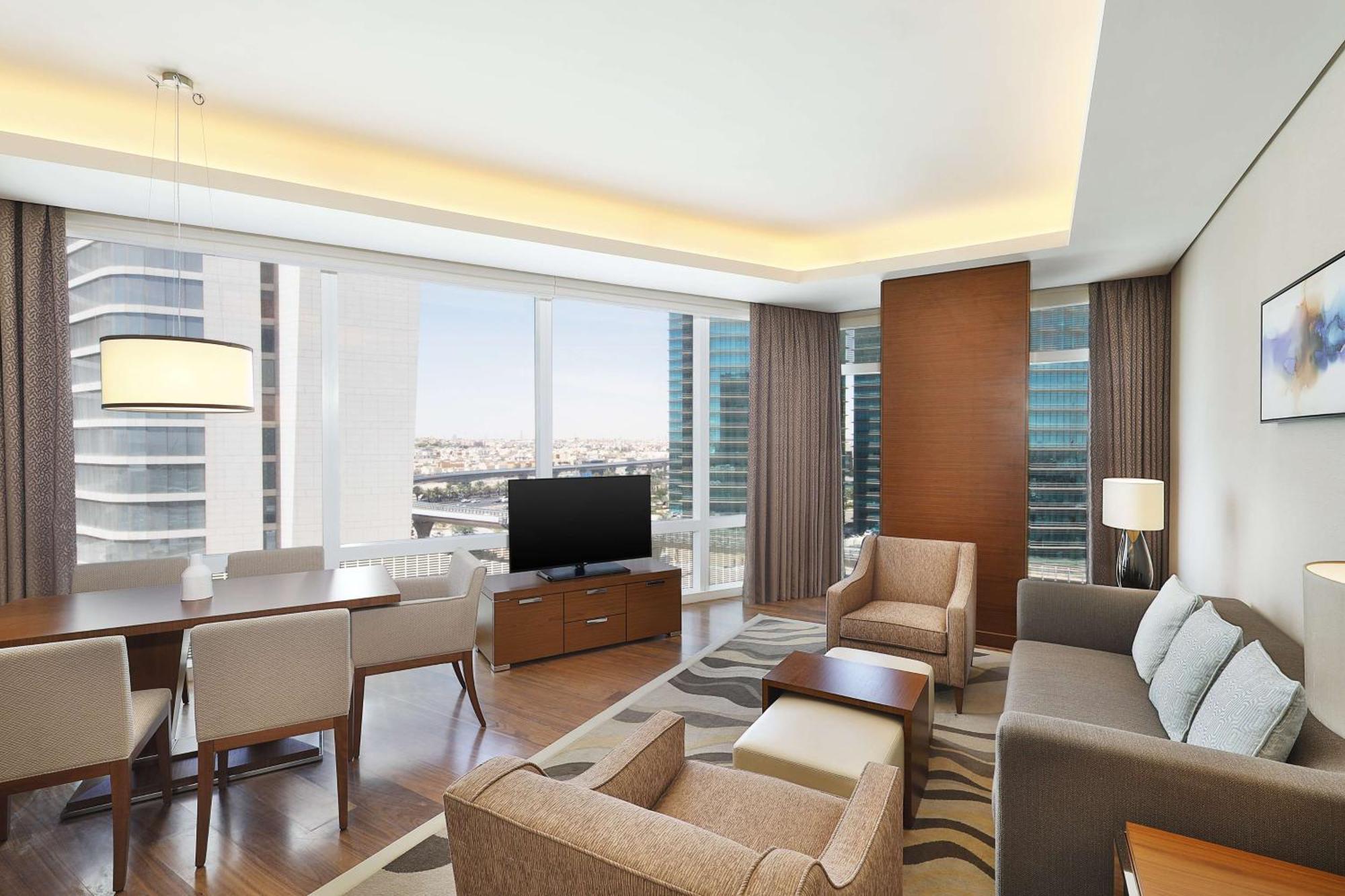 Hilton Riyadh Hotel & Residences Kültér fotó The image shows a modern, stylish living area in an apartment or hotel suite. It features large windows that provide a view of the cityscape, allowing natural light to flood the space. The furnishings include a comfortable sofa, armchairs, and a coff