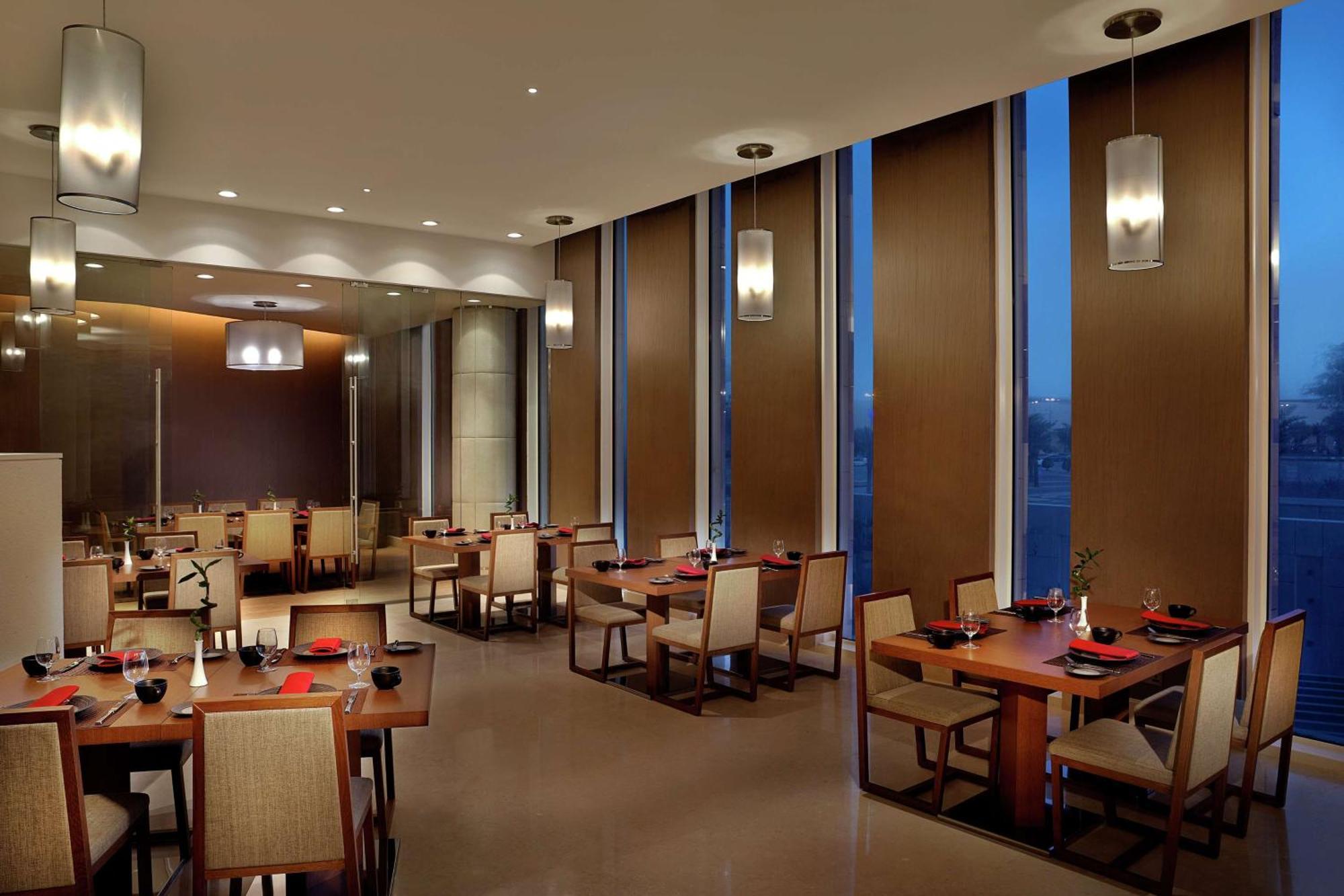 Hilton Riyadh Hotel & Residences Kültér fotó The image depicts a modern restaurant interior. It features several dining tables arranged neatly, each set with plates and utensils. The room has large windows with blinds partially drawn, allowing some ambient light to filter in, while evening ligh