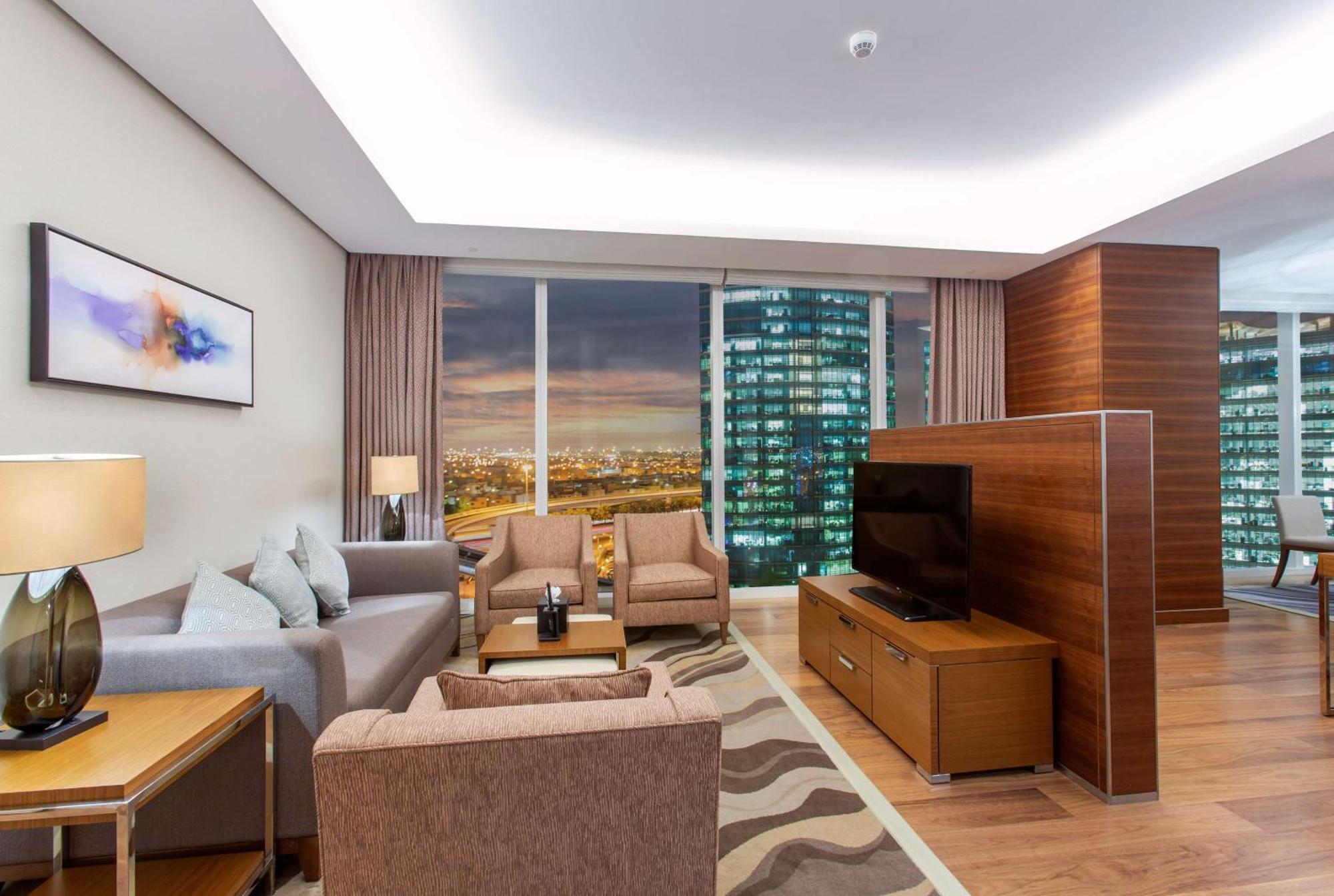 Hilton Riyadh Hotel & Residences Kültér fotó The photo shows a modern living room in a stylish apartment. The space features a comfortable seating arrangement with a light gray sofa and two brown armchairs, complemented by a wooden coffee table. There is a large flat-screen TV mounted on a wood