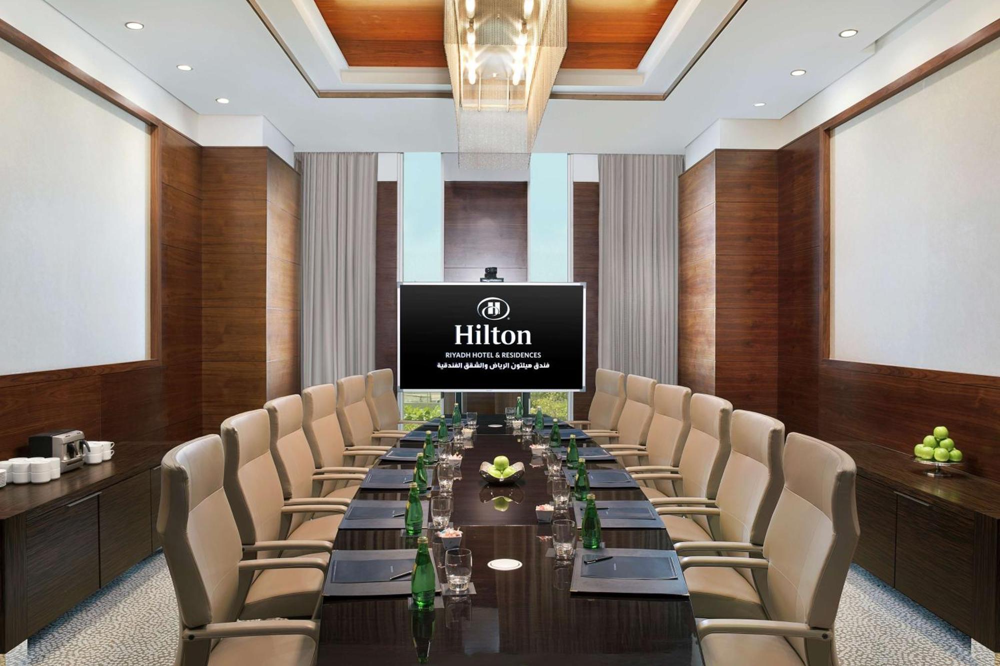 Hilton Riyadh Hotel & Residences Kültér fotó The photo shows a modern conference room within a Hilton hotel. There is a large wooden table surrounded by several comfortable chairs. At the head of the table is a large screen displaying the Hilton logo. The room is well-lit with stylish overhead 