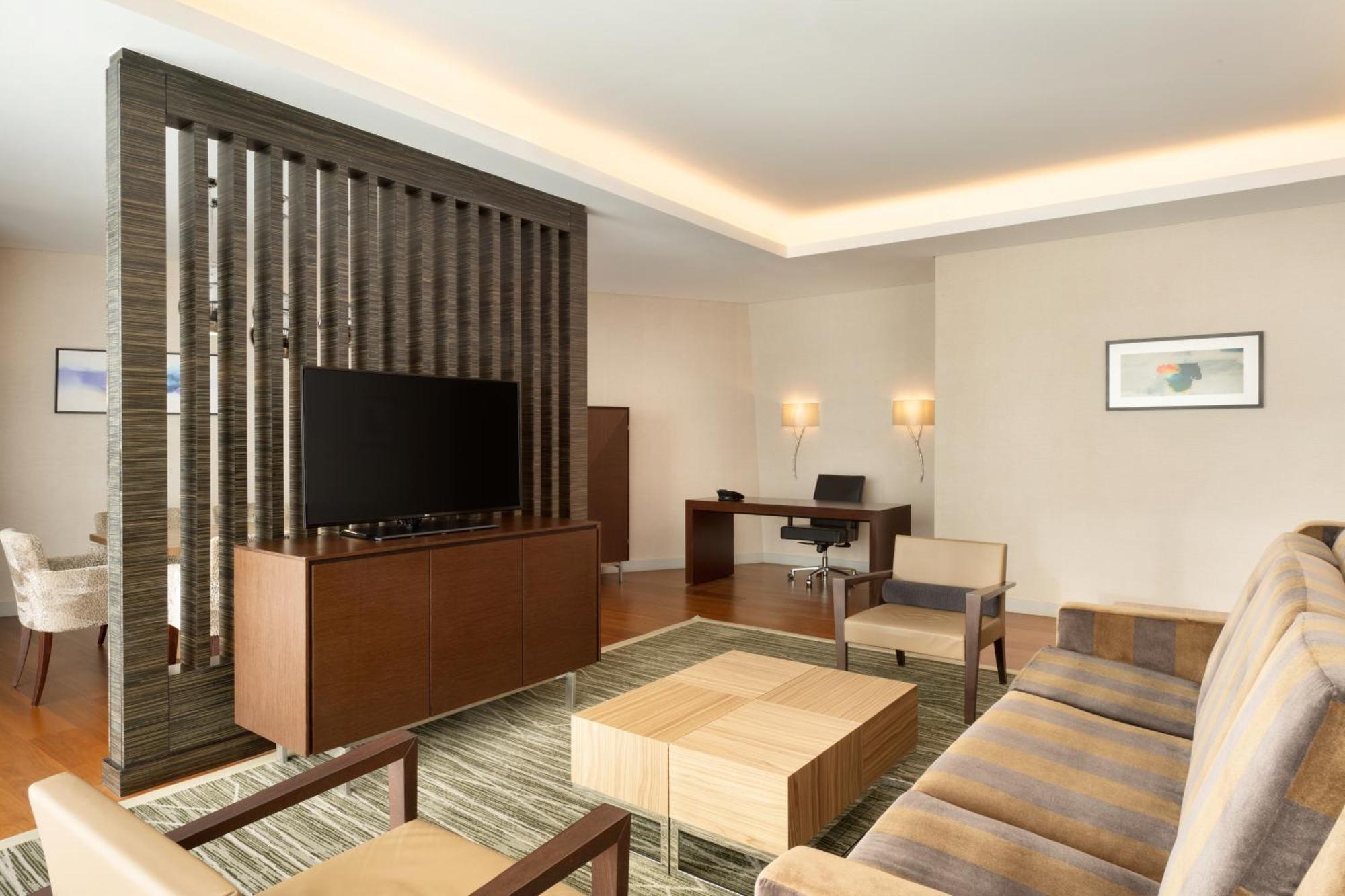 Hilton Riyadh Hotel & Residences Kültér fotó The photo shows a modern living space that features a contemporary design. It includes a seating area with a beige sofa and two armchairs arranged around a square wooden coffee table. A wall-mounted flat-screen TV is positioned on a decorative wooden
